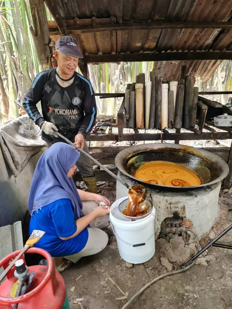 Two farmers at a wood-fired wok stove; one stirs the boiling sap while the other strains the finished sugar into a pail.
