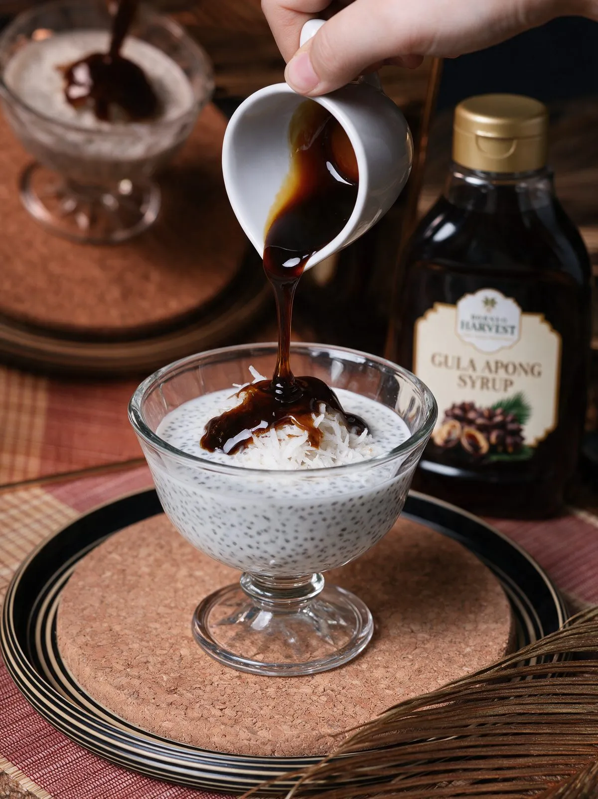 Syrup poured from a small white jug over a glass of chia seed pudding topped with shredded coconut, a bottle of Gula Apong syrup behind.