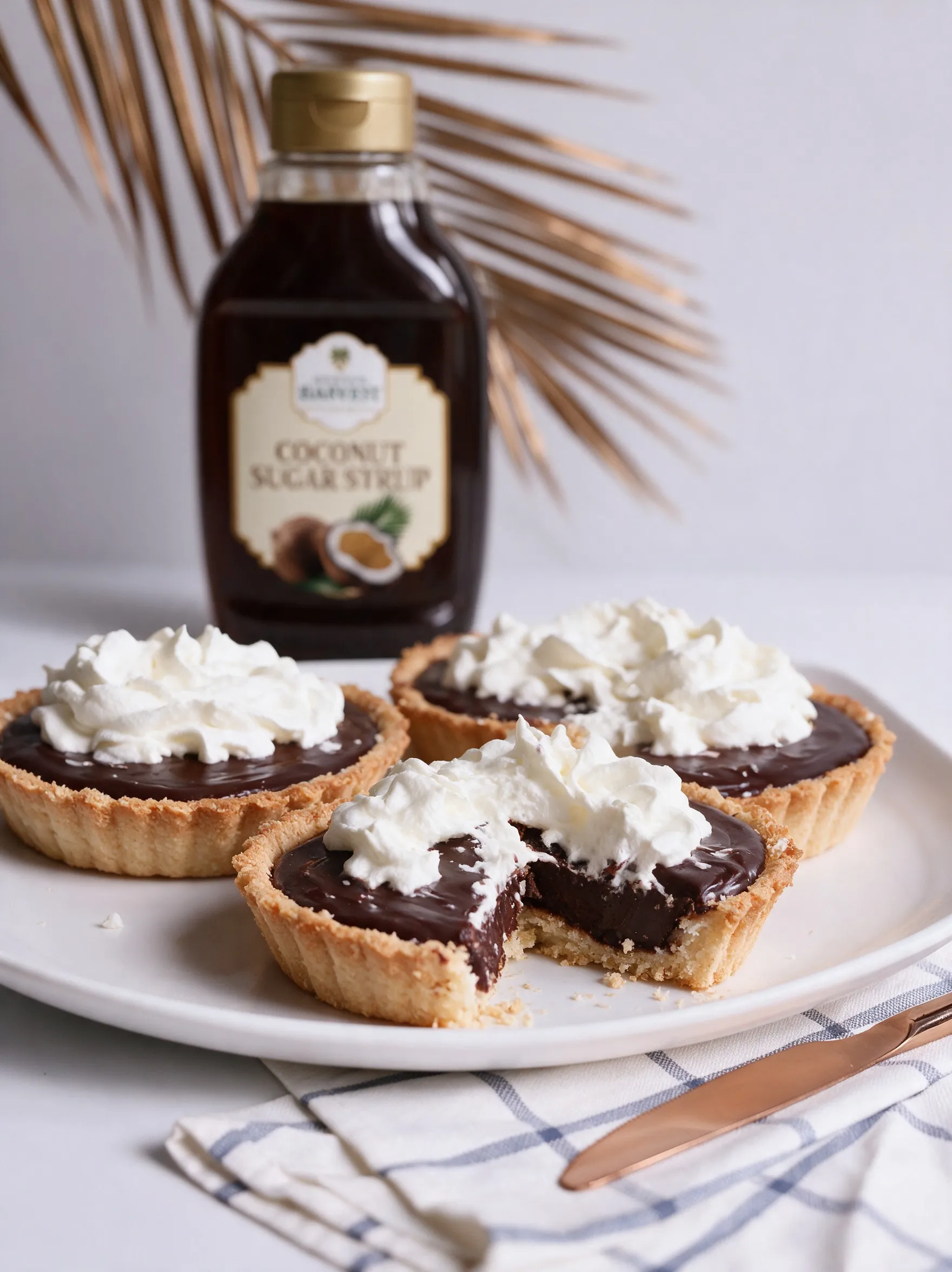 Three chocolate ganache tarts topped with whipped cream, one cut open, a bottle of coconut sugar syrup and a gold palm frond behind.