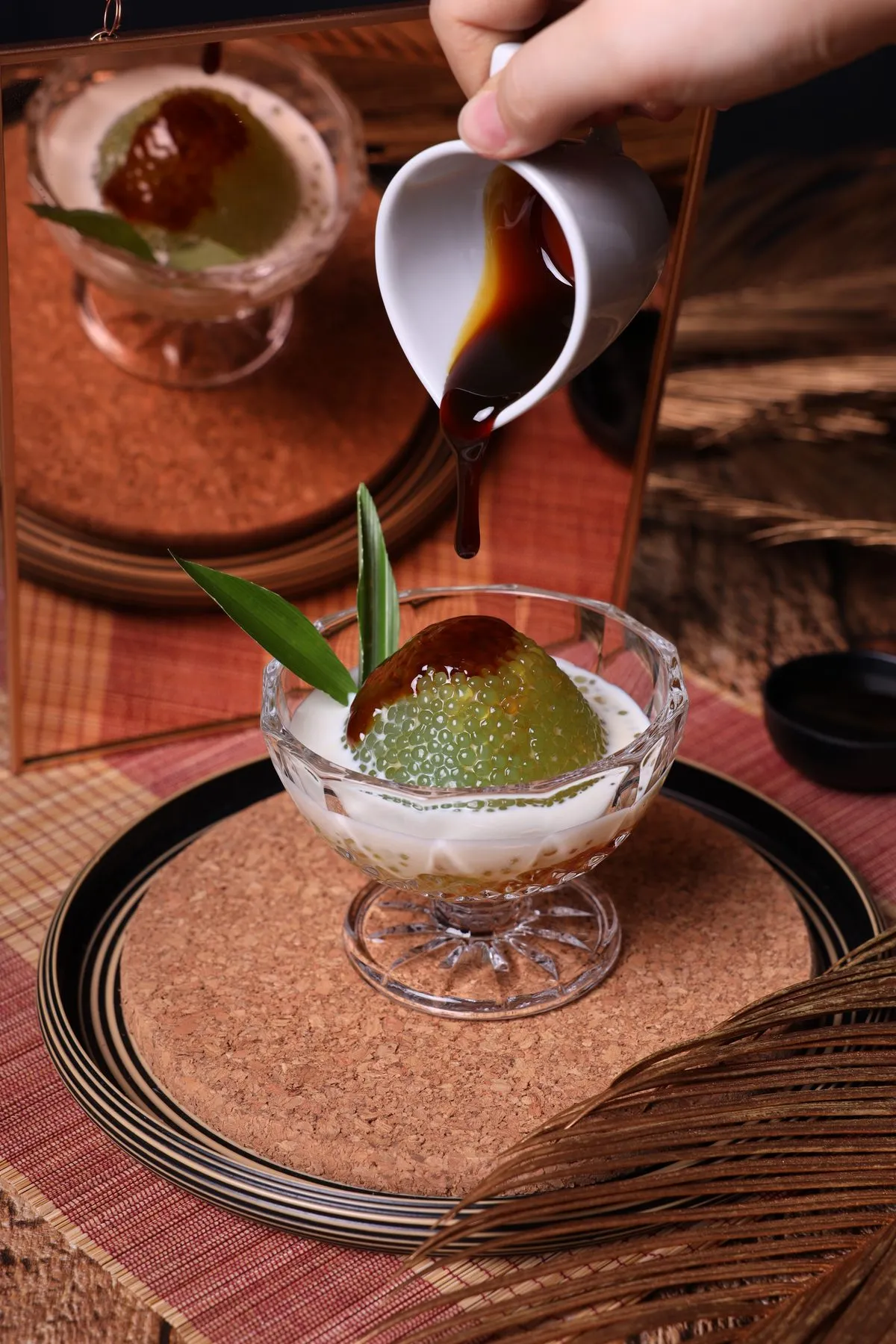 Coconut sugar syrup poured from a small white jug over a dome of pandan sago pearls sitting in coconut milk in a glass dish, a pandan leaf tucked in beside it and the dish reflected in a mirror behind.
