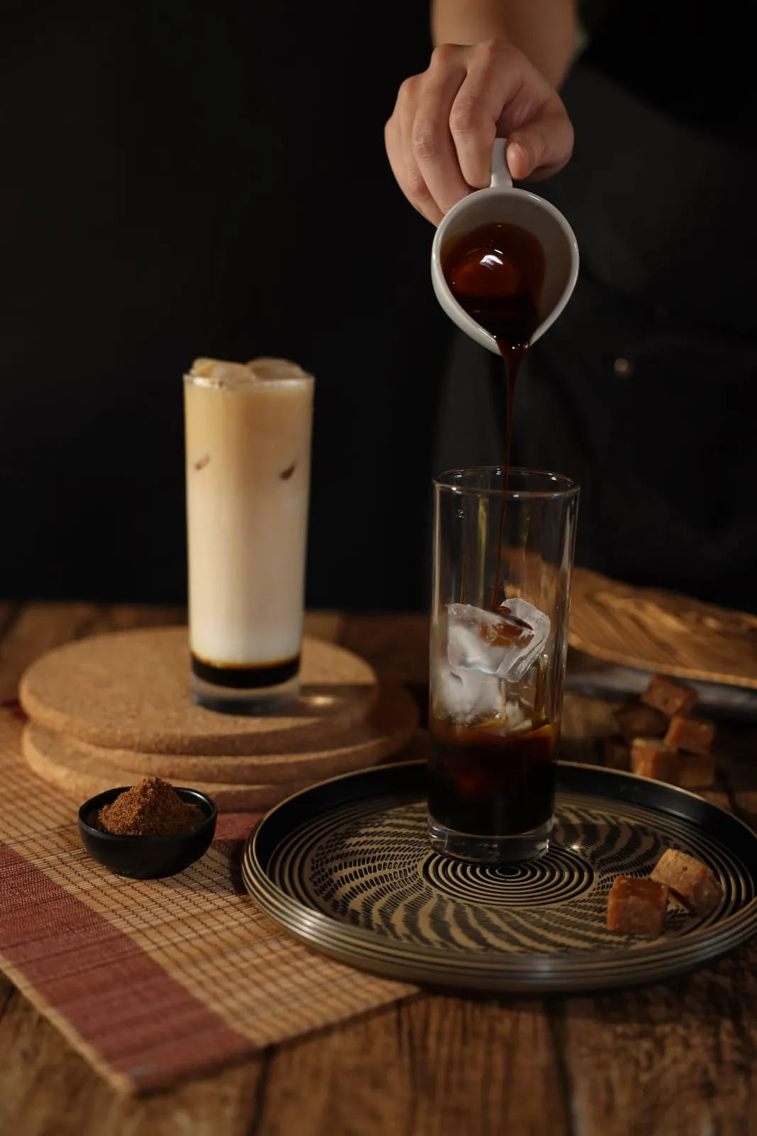 Gula Apong syrup poured from a white jug over ice in a tall glass, a finished three-layer milk tea standing behind.