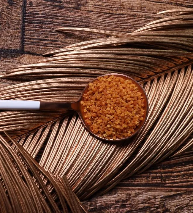 A copper spoon of golden coconut sugar granules resting on a gilded palm frond